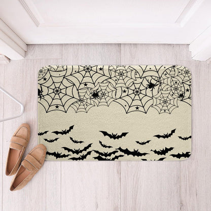 A rectangular doormat with a black spiderweb and bat pattern on a light beige background, placed on a light wood floor next to a pair of tan slip-on shoes.