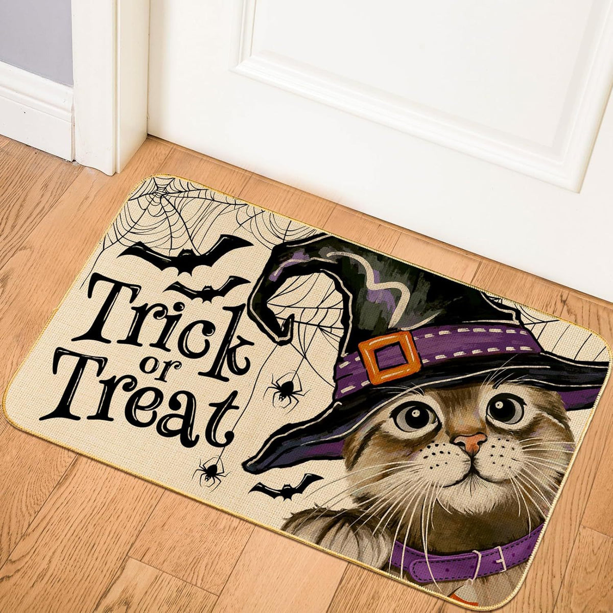 A rectangular Halloween doormat placed on a wooden floor in front of a white door. The mat features a cat wearing a witch hat, spiders, bats, cobwebs, and the words “Trick or Treat” in large black lettering.
