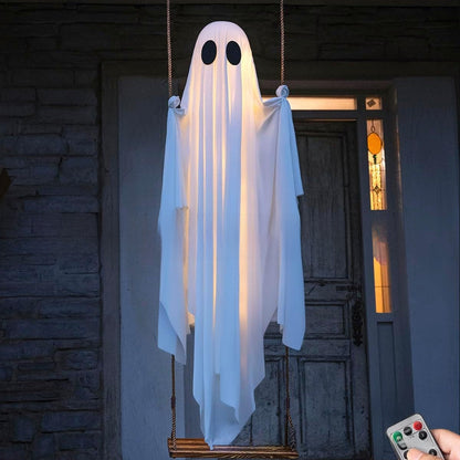 A hanging ghost Halloween decoration with black oval eyes and a white draped body, suspended in front of a dark doorway. A hand holding a remote control is visible in the lower right corner.