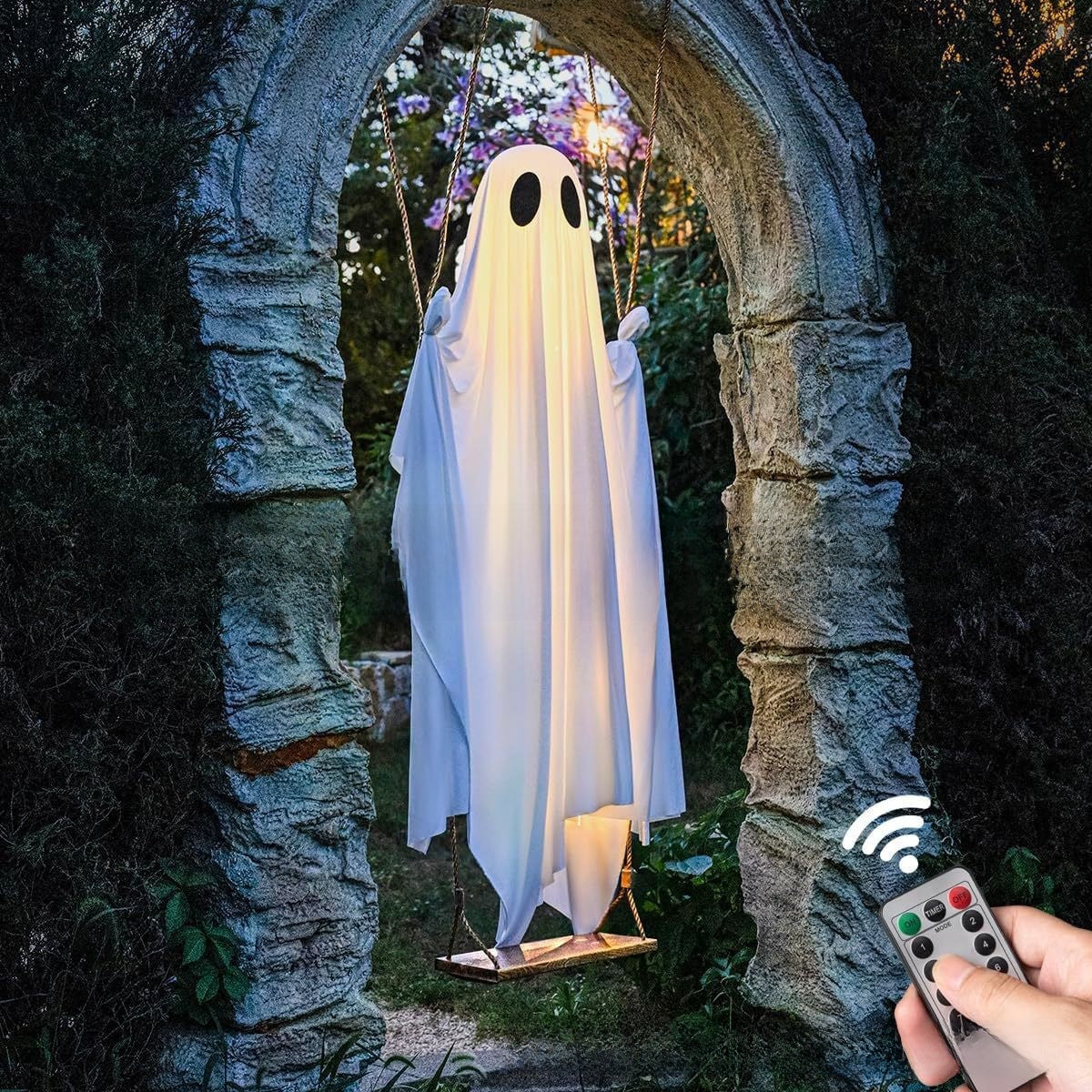 A ghost-shaped hanging swing decoration with a white sheet and black eyes, suspended in front of an arched stone doorway. A hand holds a remote control in the lower right corner, with a wireless signal graphic above it.