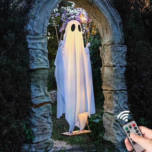 A ghost-shaped hanging swing decoration with a white sheet and black eyes, suspended in front of an arched stone doorway. A hand holds a remote control in the lower right corner, with a wireless signal graphic above it.