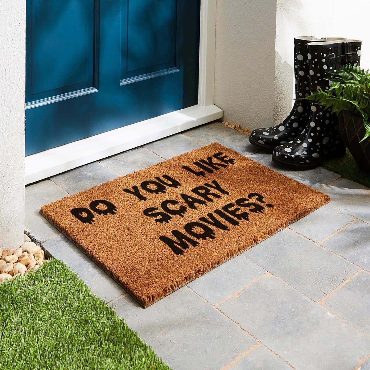 A brown coir doormat with the text 'DO YOU LIKE SCARY MOVIES?' in black, placed on a stone walkway in front of a blue door. A pair of black rain boots and a potted plant sit to the right.
