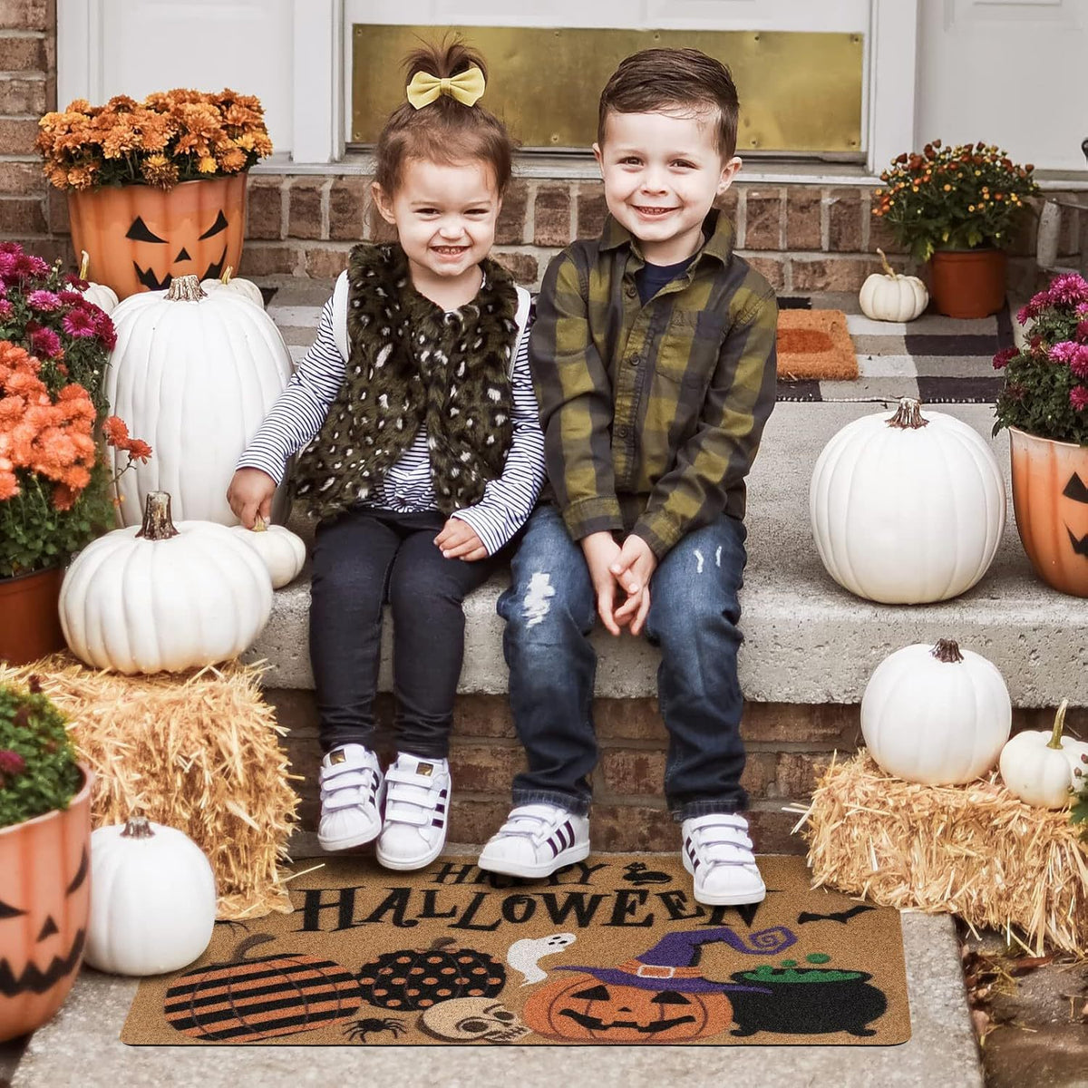 A young boy and girl sit on concrete steps in front of a house, surrounded by pumpkins, flowers, and a Halloween-themed doormat. The girl wears a brown patterned vest over a gray-and-white striped shirt and dark pants, and the boy wears a green plaid shirt and blue jeans.