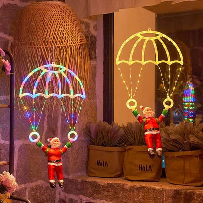 Two small Santa Claus figurines hanging from glowing parachute-shaped LED light decorations, displayed on a stone ledge with potted plants and a woven basket in the background.