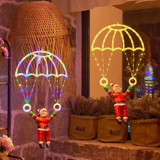 Two small Santa Claus figurines hanging from glowing parachute-shaped LED light decorations, displayed on a stone ledge with potted plants and a woven basket in the background.