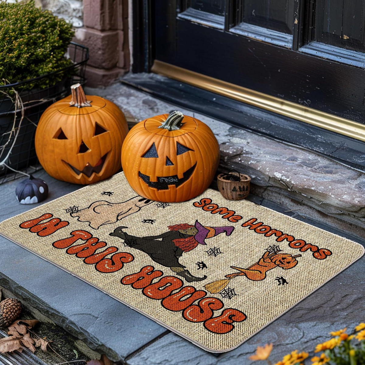 A Halloween-themed doormat with the phrase 'Some Horrors in this House' lies on a stone doorstep. Two carved pumpkins, a small black cat figurine, and a small basket sit beside the mat, with a dark door and plants in the background.