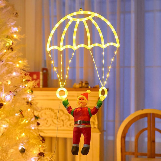 A festive Christmas hanging ornament shaped like an elf dangling from a glowing yellow parachute with string lights, set indoors near a lit Christmas tree and window.