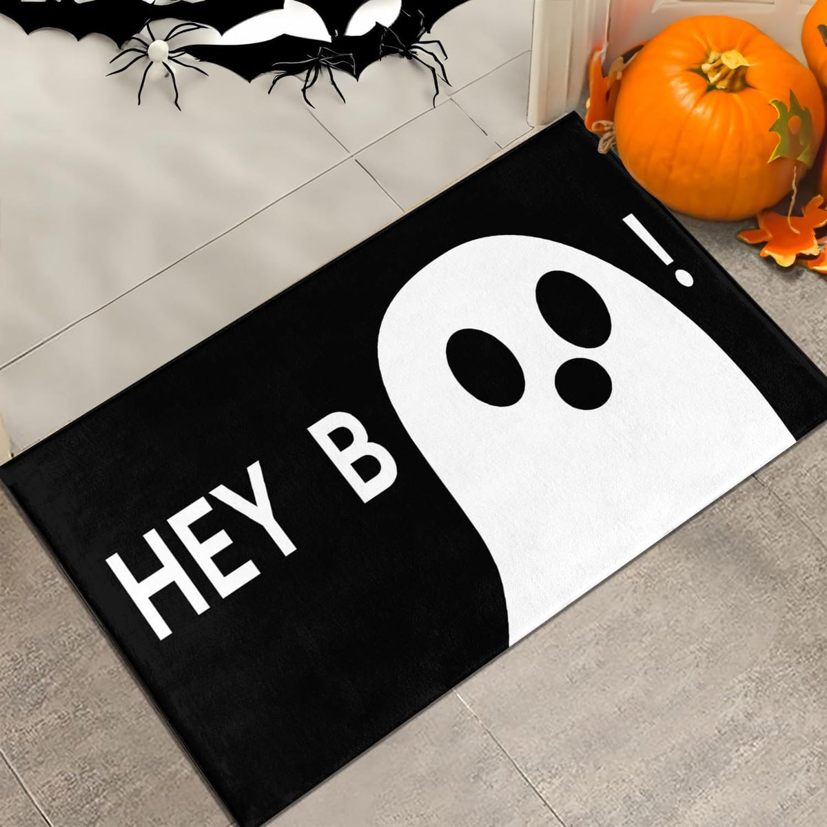 A black and white Halloween doormat with a ghost graphic and the text 'HEY B', shown on a gray tiled floor with pumpkins and spider decorations nearby.
