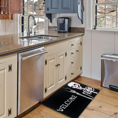 A black kitchen floor mat with a white skeleton graphic and the word 'WELCOME' in white, placed on a wood floor in a kitchen with cream cabinets and stainless steel appliances.