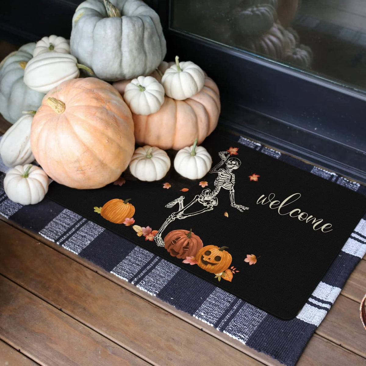 A black fall-themed doormat with a skeleton, pumpkins, leaves, and the word 'welcome' in script, styled on a wooden porch with decorative pumpkins around it.