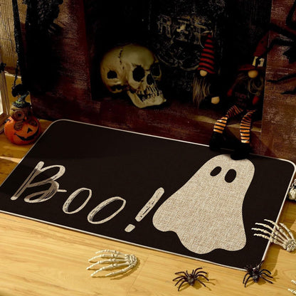 A Halloween-themed doormat with 'Boo!' in script and a ghost graphic, displayed on a wooden floor surrounded by spooky props including a skull, gnomes, spiders, and skeleton hands.
