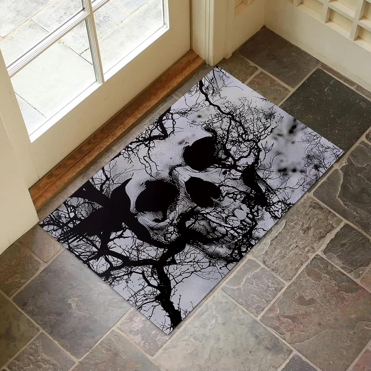 A rectangular black and white doormat featuring a skull and bare tree branches design, placed on a stone tile floor in front of a glass door.