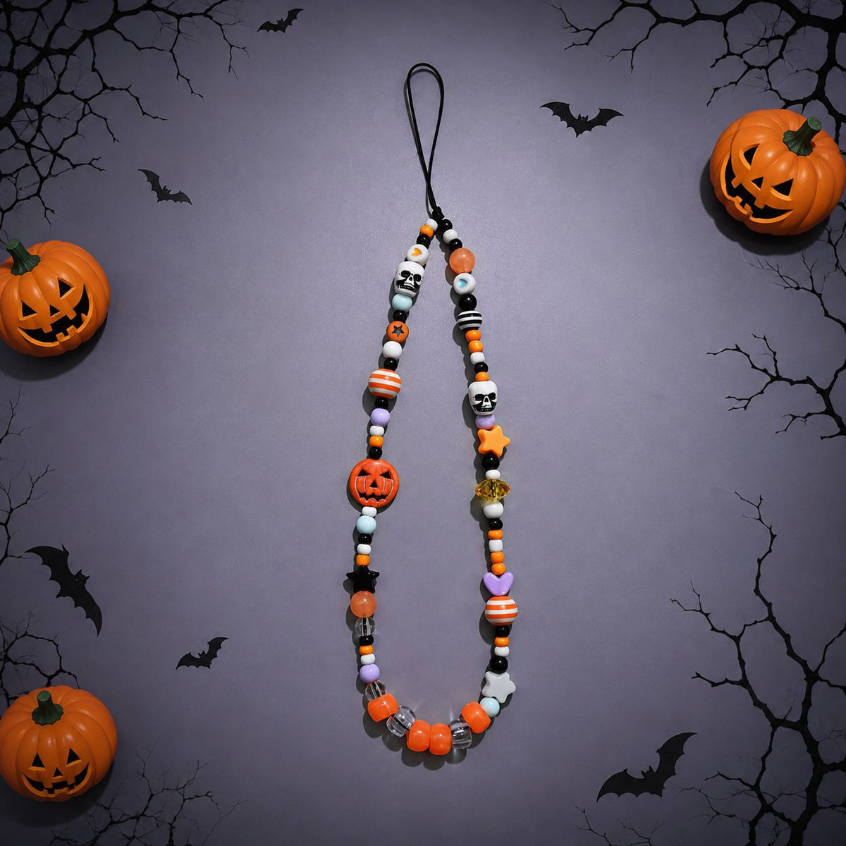 A Halloween-themed beaded necklace or phone strap featuring orange, black, white, and purple beads shaped like pumpkins, skulls, bats, and candy canes, displayed against a dark gray background with decorative jack-o'-lanterns and black tree branches.
