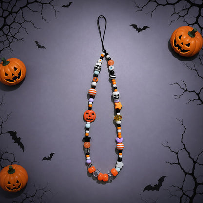 A Halloween-themed beaded necklace or phone strap featuring orange, black, white, and purple beads shaped like pumpkins, skulls, bats, and candy canes, displayed against a dark gray background with decorative jack-o'-lanterns and black tree branches.