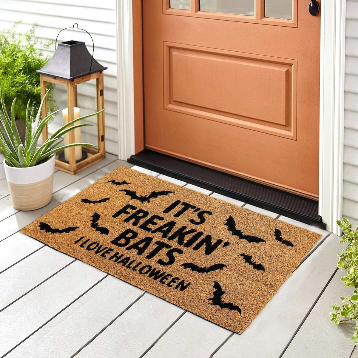 A rectangular coir doormat with black text reading 'IT'S FREAKIN' BATS I LOVE HALLOWEEN' and black bat silhouettes, placed on a light porch in front of a brown front door. A lantern, potted plant, and house siding are visible around it.