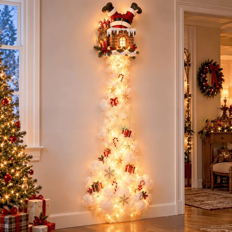 A festive Christmas wall decoration shaped like Santa's legs stretching down into a fluffy white cloud-like base, decorated with warm white lights, ornaments, candy canes, snowflakes, and a gingerbread house at the top, shown in a decorated home hallway.