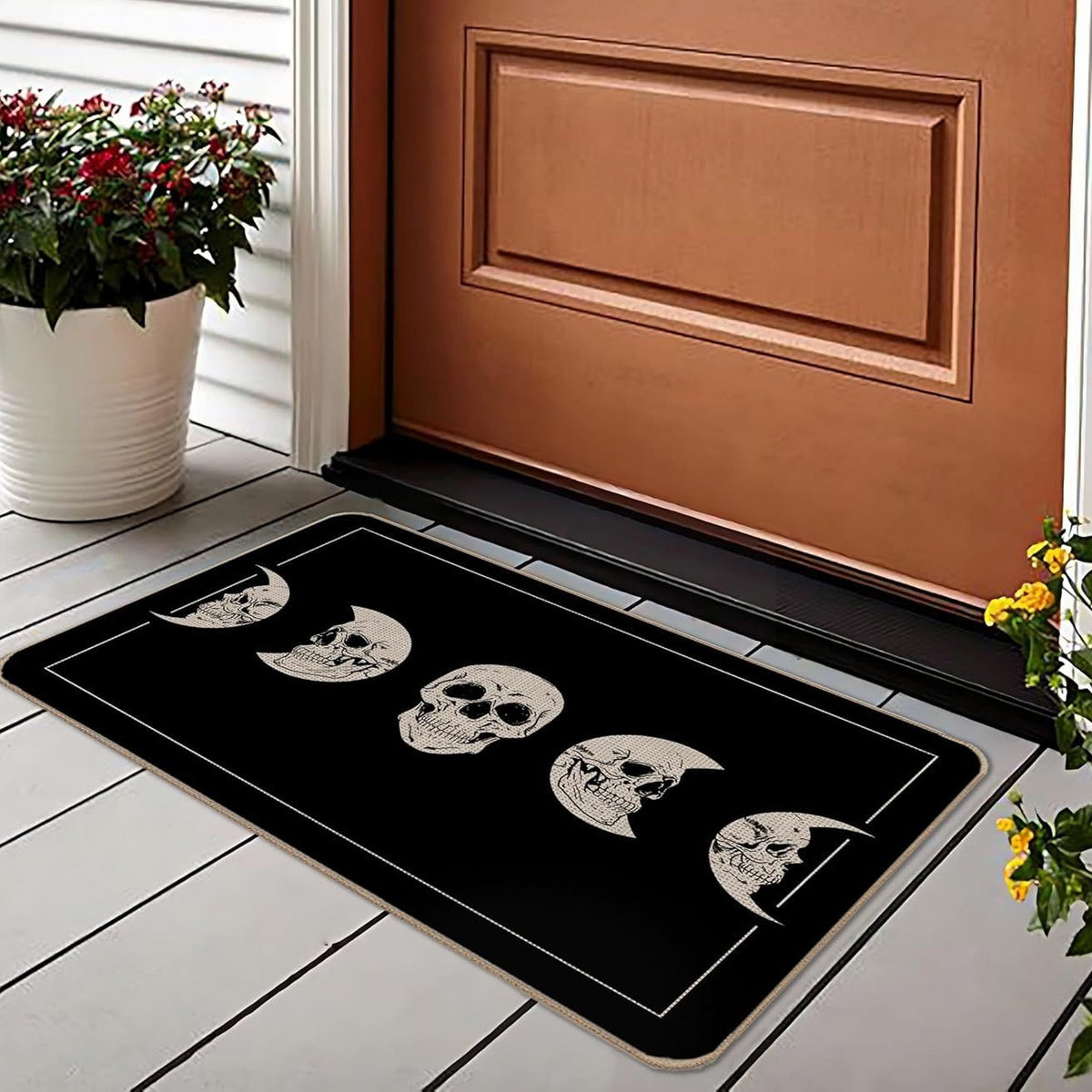 A rectangular black doormat with a row of white skull and moon phases printed across it, placed in front of a brown door on a white tiled porch with potted plants nearby.
