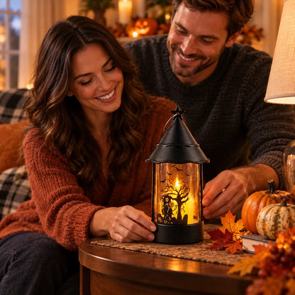 A couple smiling in a cozy living room setting, looking at a black metal Halloween lantern with a warm glowing tree silhouette on a round wooden table decorated with autumn leaves and pumpkins.