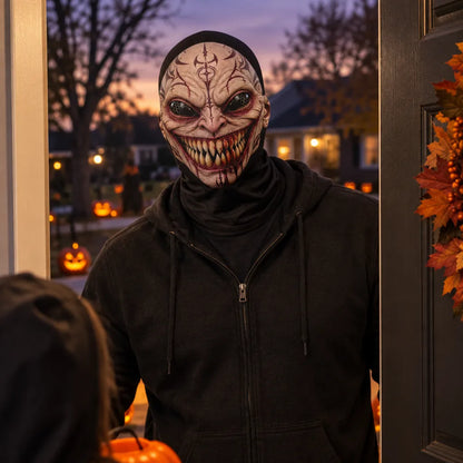A person wearing a scary, horror-style mask and a black zip-up hoodie stands in a front door at dusk, with pumpkins and a decorated wreath visible in the background.